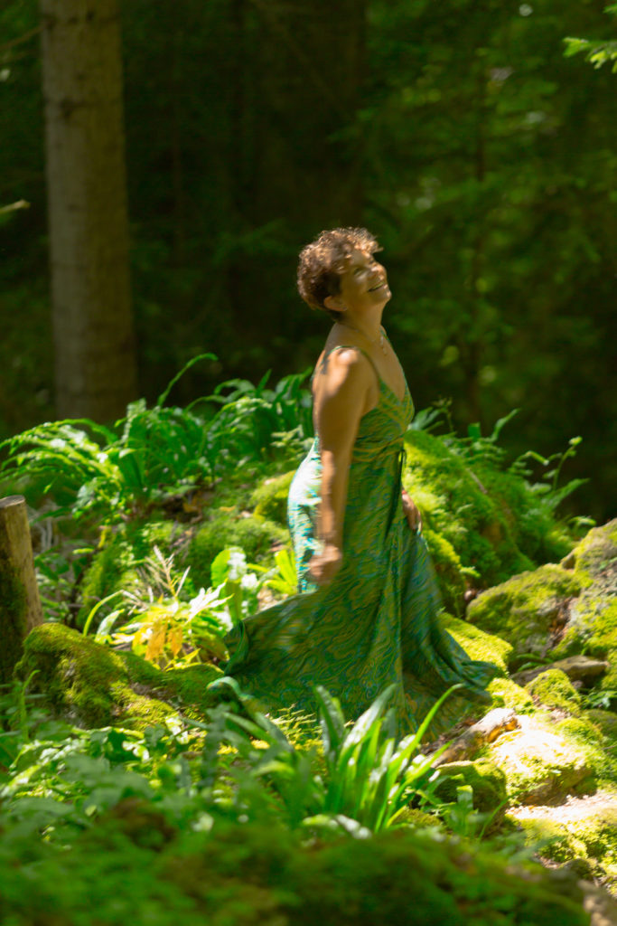 Portrait de Véronique Wampfler souriante en robe turquoise dans une forêt luxuriante et mousseuse. Ambiance féérique et lumineuse.